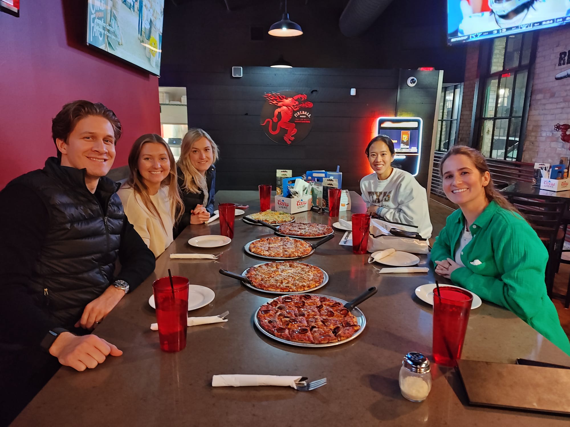 five people sitting around a rectangular table with five pizzas on the table