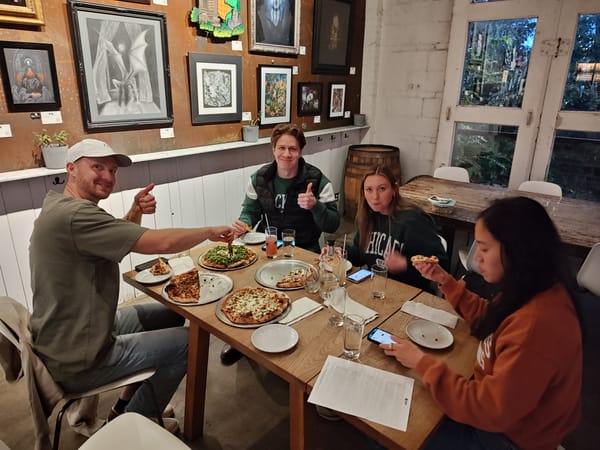 four people smiling and sitting at a table with four pizzas on the table